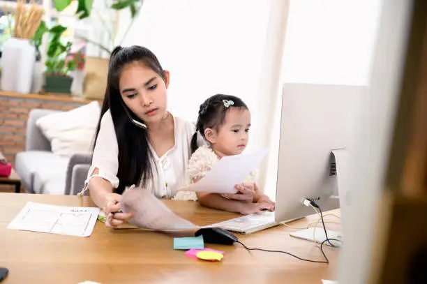 Photo of Beautiful middle aged Asian woman or businesswoman working on her cell phone and laptop computer at home and take care her baby daughter.