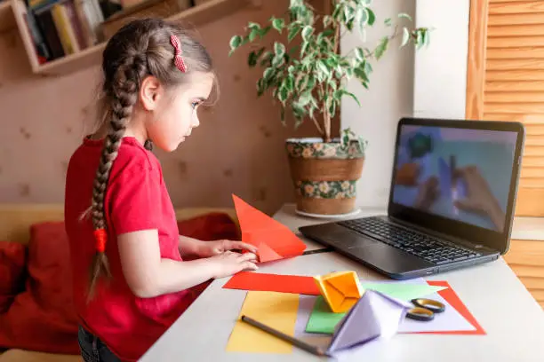 Cute little elementary schoolgirl doing origami fish with folded color paper looking video on laptop, online workshop, kids at-home activity, creativity and distant education