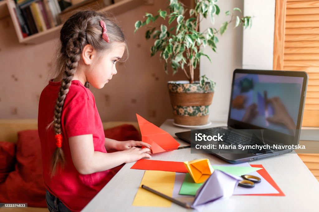 Flicka gör origami fisk med färgpapper söker video på laptop, online verkstad och avlägsen utbildning - Royaltyfri Barn Bildbanksbilder