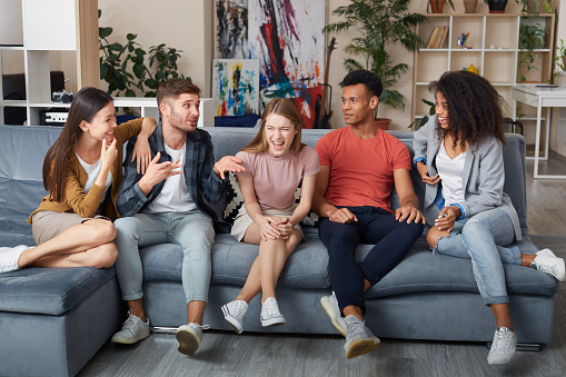 Spending nice time together. Group of young happy multicultural people in casual wear laughing while sitting on the sofa at home. They relaxing together in the modern apartment. Home party. Friendship
