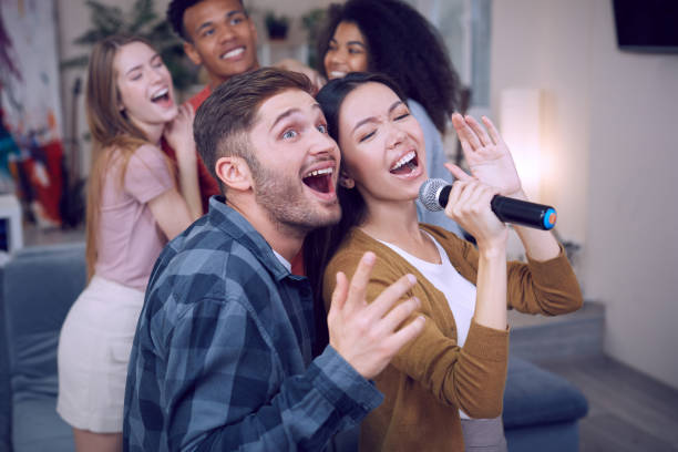 istock Singing duet. Young excited couple or friends holding microphone and singing together while playing karaoke with friends at home. Group of people having fun, karaoke party 1222375863