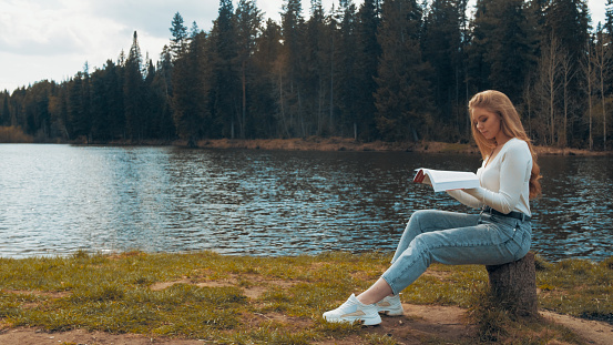 Beautiful young blonde woman sitting and reading book in a park near the lake. Side view.