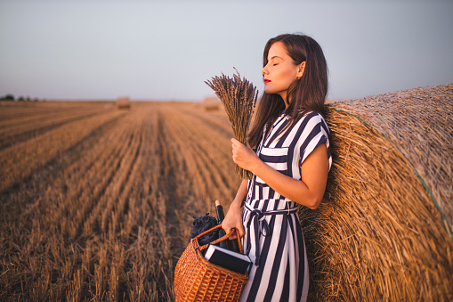 Fashion woman holding picnic basket and standing on harvested field with straw bales