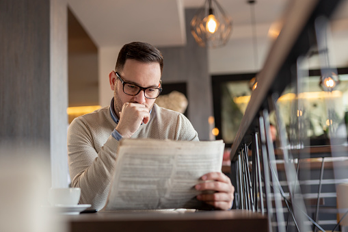 Pensive man sitting at a restaurant table, reading newspaper and drinking coffee