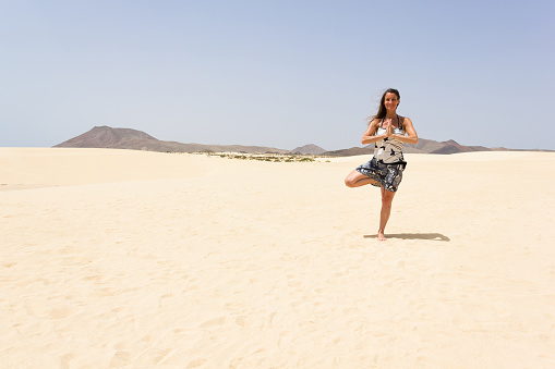 Middle aged woman practicing tree yoga balance pose on sand dunes in Fuerteventura island, Spain. Hope, metaphor concepts