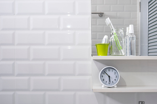 Toothbrush in glass bottle, tooth paste in little green bucket, hand sanitizer spray with clock on shelf and mirror on white tiles wall in modern bathroom, personal accessories