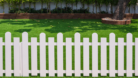Front view of white wooden fence in front of artificial turf with green plant growing on interlocking brick blocks in front yard of home