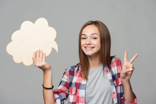 feliz chica adolescente sosteniendo burbuja de pensamiento y gesturing v signo - decisions teenage girls horizontal studio shot fotografías e imágenes de stock