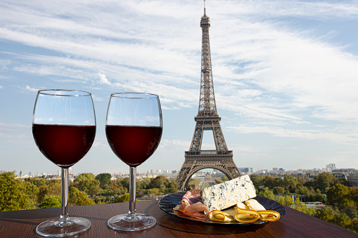 Two glasses of red wine with charcuterie assortment on view of Paris and Eiffel tower background. Glass of red wine with different snacks - plate with ham, sliced cheese, blue cheese. Couple romantic celebration in Paris.