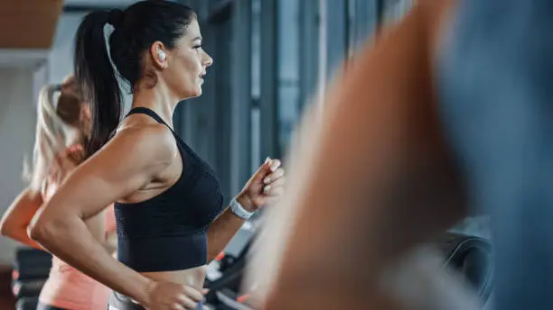 Beautiful Athletic Sports Woman Wearing Wireless Headphones, Listens to a Podcast or Sport Music Playlist while Running on a Treadmill. In Background Fit Athletes Training in the Gym