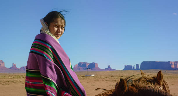 ein teenaged native american girl in einer traditionellen navajo decke eingewickelt sitzt auf ihrem pferd und schaut auf die kamera in der monument valley wüste mit großen felsformationen in der ferne an einem klaren, hellen tag - monument valley navajo mesa monument valley tribal park stock-fotos und bilder