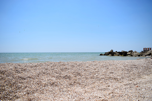 Bridge to the sea. Rocks on the water. Glare on the blue sea water. Sand and shells on the beach.