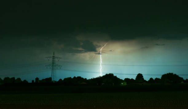 Dangerous severe storm approaches high voltage electricity power lines in agricultural field: steel pylons carrying multiple power cables appear to stand defiant as the maelstrom bears down. Wide angle view