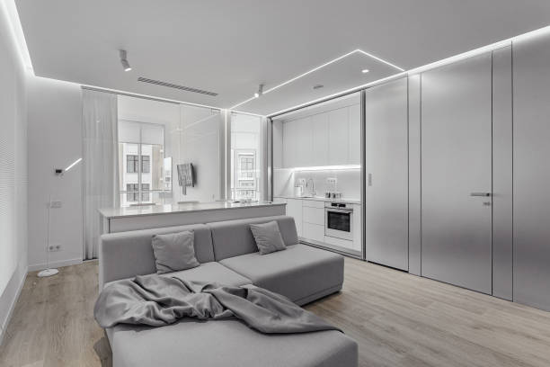 istock Spacious white guest area, gray sofa with drapery, two pillows. compact stylish kitchen and bedroom behind glass door. Panoramic windows. Lots of air and space 1222435280