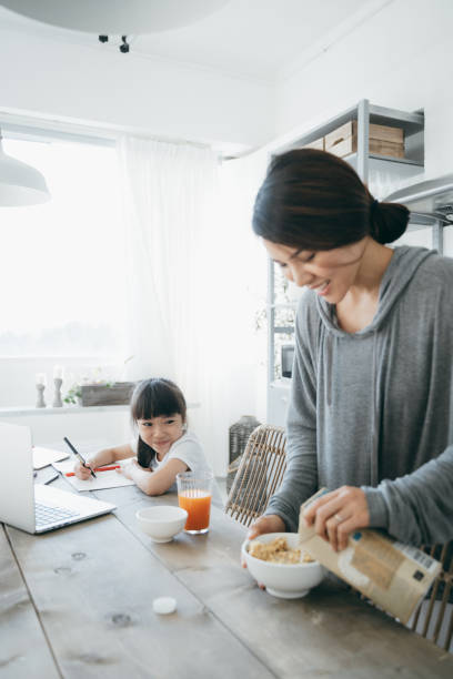 istock Young Asian mother preparing healthy breakfast while little daughter is studying from home. She is attending online school classes with a laptop and writing notes at home 1222436977