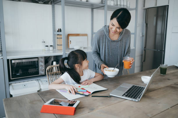 istock Young Asian mother serving healthy breakfast on the table while little daughter is studying from home. She is attending online school classes with a laptop and writing notes at home 1222437000