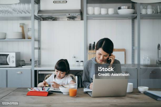 Young Asian Mother Working From Home On A Laptop And Talking On The Phone While Little Daughter Is Studying From Home She Is Attending Online School Classes With A Digital Tablet And Doing Homework At Home Stock Photo - Download Image Now