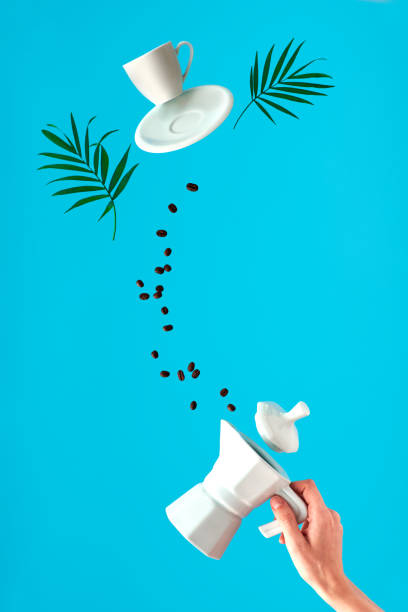 Ceramic stove coffee maker in woman's hand. Trendy levitation. Flying coffee beans and espresso cup with saucer. Blue mint background with palm leaves, text space.