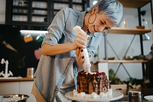 Happy woman decorating homemade cake with whipped cream at home kitchen