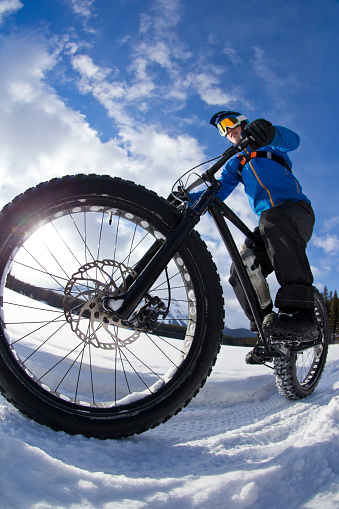 A close-up view of a young man on a winter fat bike ride along the edge of a frozen lake in the Rocky Mountains of Canada. Fat bikes are mountain bikes with oversized wheels and tires for riding on the snow. He wears a bicycle helmet, goggles, carries a backpack, and wears warm winter clothing.