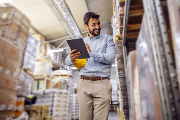 toda la longitud de apuesto hombre de negocios sonriente que tiene una llamada telefónica con el cliente mientras camina en el almacén. - document shipping freight transportation form fotografías e imágenes de stock