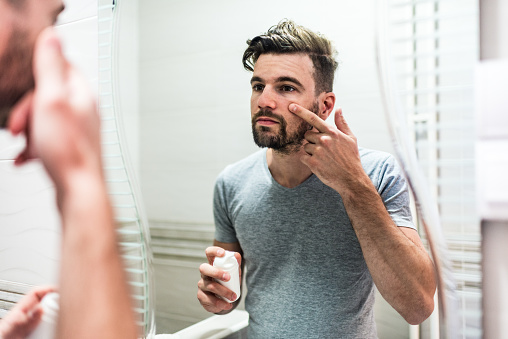 Young man taking care of his skin. Man applying skincare facial treatment cream on face in the bathroom. Handsome guy applying moisturizer and looking at himself while standing in front of the mirror.