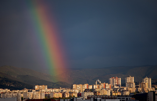 Rainbow raising above city buildings in rainy weather just before sunset