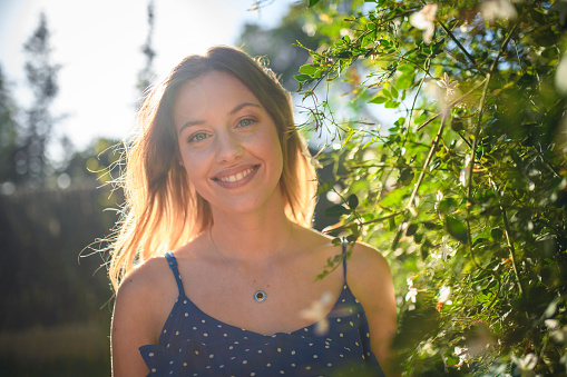 Front view with lens flare of relaxed young woman in casual summertime clothing standing next to green foliage and smiling at camera.