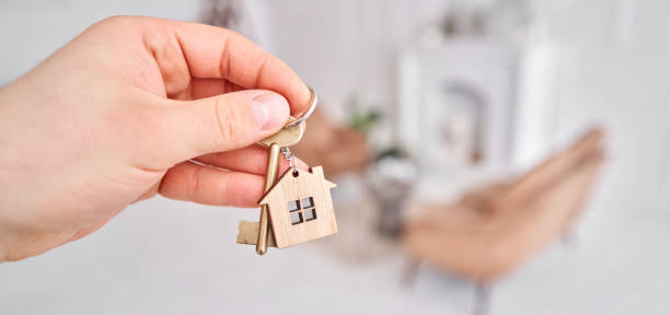 istock Men hand holding key with house shaped keychain. Modern light lobby interior. Mortgage concept. Real estate, moving home or renting property. 1222520882