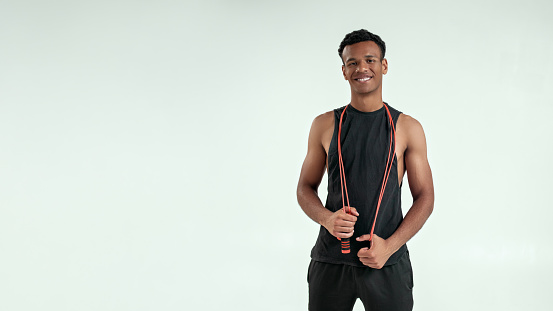 Best cardio. Strong and healthy young afro american man holding a jumping rope at his shoulders and looking at camera with smile while standing against grey background. Sport equipment. Active lifestyle