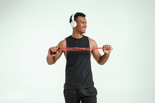 Exercising with music. Cheerful and sporty young afro american man in headphones holding a jumping rope and smiling while standing against grey background. Sport equipment. Active lifestyle