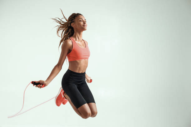 Flying. Happy and young african woman with perfect body skipping rope while exercising in studio against grey background stock photo