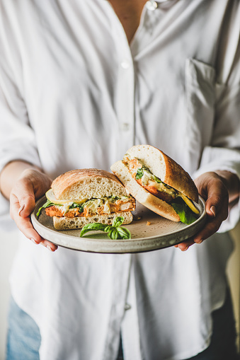 Woman in white shirt holding plate with fresh fried fish sandwich with tartare sauce, lemon and arugula cut in halves, selective focus. Healthy easy breakfast ideas