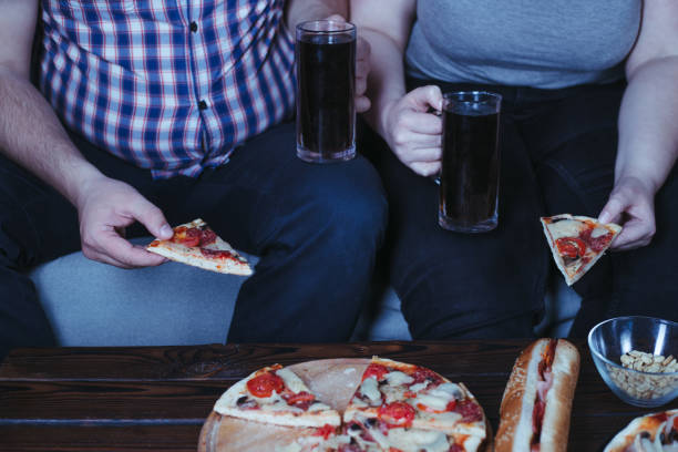 coppia in sovrappeso che guarda la tv mangiare la pizza - abdomen addiction adult alcohol foto e immagini stock