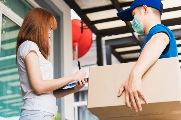 Asian young delivery man courier shopping online with package post box in uniform protective face mask coronavirus he making service woman customer using pen signature on paper book at home, COVID-19 asiático joven repartidor mensajero comprando en línea con el paquete de correos caja en uniforme protector máscara facial coronavirus que hace cliente de la mujer de servicio utilizando la firma de la pluma en el libro de papel en casa, covid-19