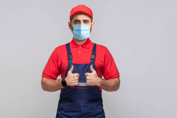 il lavoro è finito! ritratto di giovane con maschera medica chirurgica in tuta blu, maglietta rossa, berretto, in piedi e mostrando tonf e guardando la macchina fotografica. - thumps up foto e immagini stock