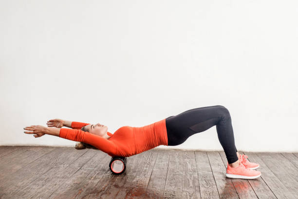 mujer delgada en pantalones ajustados deporte haciendo ejercicio con masajeador de rodilloespuma en el suelo, relajando y estirando los músculos de la columna vertebral - warming up fotos fotografías e imágenes de stock