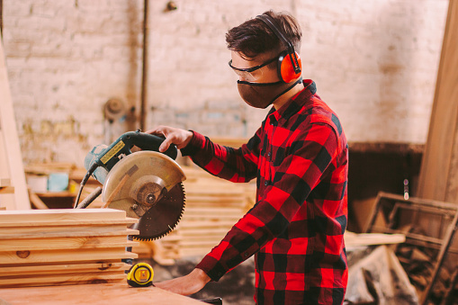 Carpenter in protective glasses, headphones and mask using electric circular saw at sawmill. Professional skilled cabinet maker cutting wood board with circle saw. Woodworking, furniture production