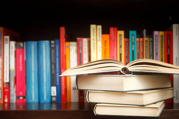 Row of books on a shelf, multicolored book spines, stack in the foreground.. Stack of books on a wooden library shelf, the one on top open , multicolored book spines in a row in the background. Copy space on the left. book stock pictures, royalty-free photos & images