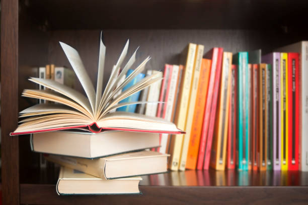 Row of books on a shelf, multicolored book spines, one open in fan shape. Row of books on a wooden library shelf, multicolored book spines, stack in the foreground ,the one on top open in fan shape. Copy space on the right. paperback-book-stack-book-spine stock pictures, royalty-free photos & images
