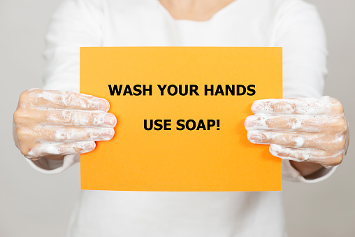 Unrecognizable female is holding a white paper to camera with soapy hands. There is wash your hands for 20 seconds written on one the paper referring to hygiene rules of pandemic conditions.