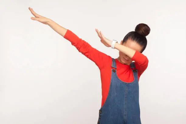 Photo of Winning, success gesture. Portrait of stylish girl with hair bun in denim overalls showing dab dance pose, famous internet meme
