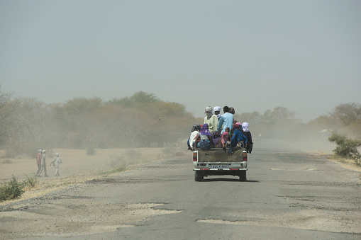 Bitkine, Chad - February 20,2020: A group of people from rural Chad is travelling on a pick-up car. The air is full of sand from the Sahel zone and the close-by Sahara desert. The scene happens is a street in the small town of Bitkine, which is an important trading place in Central Chad.