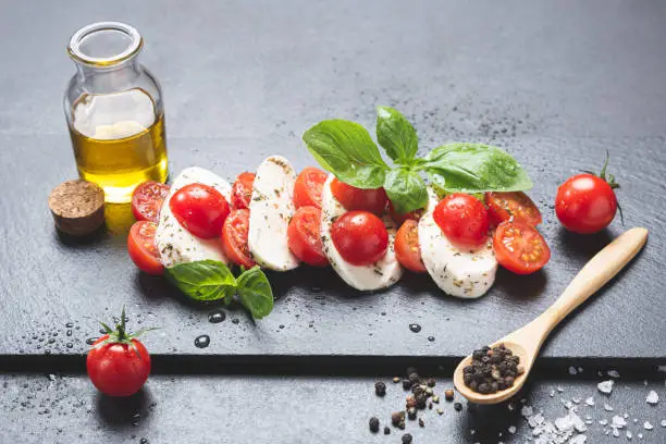 Mozzareela, tomato and basil, on blackboard on black background, 
with extra virgin olive oil, salt and pepper. 
Mediterranean diet 
and healthy food concept