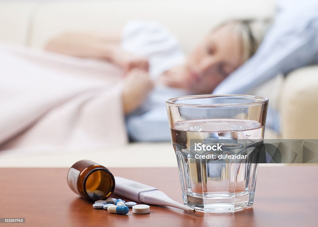 Medicine, Thermometer, and Water Left out for a Sick Woman Pills, thermometer and water with girl in bed in background Adult Stock Photo