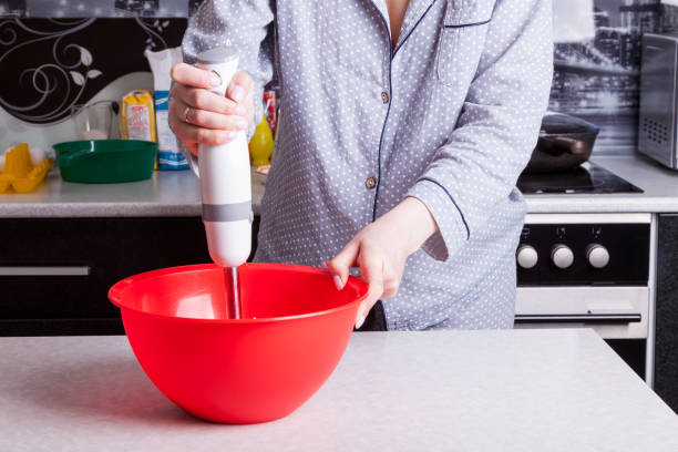 girl in pajamas in the kitchen prepares breakfast holding a hand blender in her hand and mixes ingredients for omelet or pancakes for the whole family. - cake making mixing eggs imagens e fotografias de stock