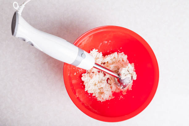 submersible blender with a light handle and a metal nozzle in a red bowl with mixed curd and sugar for making cheesecakes for breakfast close-up isolated on a white background. - cake making mixing eggs imagens e fotografias de stock