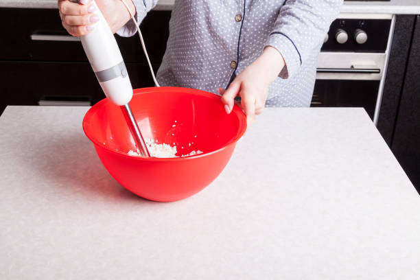 руки женщины держат подводный блендер и смешивают ингредиенты для теста и панеттоне дома в красной миске на белом кухонном столе. хобби вып - cake making mixing eggs стоковые фото и изображения