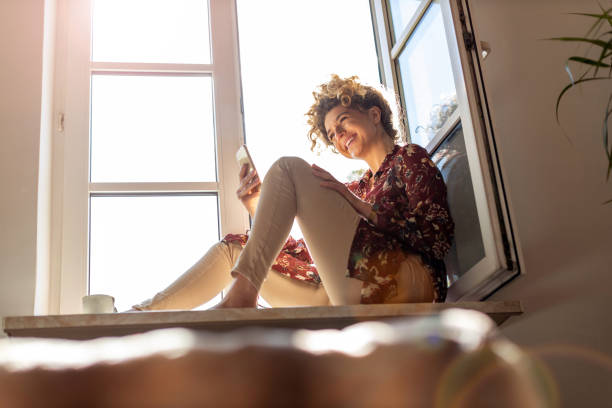 istock Young woman sitting on window sill and using smartphone 1222587071