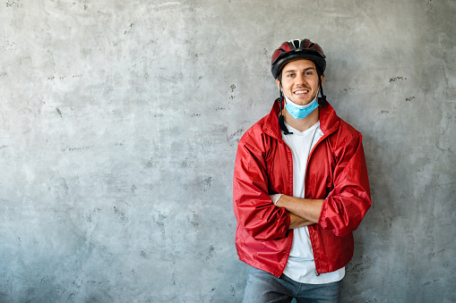 Young diligent male delivery person wearing protective face mask and cycling helmet, as he proudly standing with arms crossed
